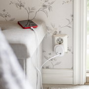 Weißes USB-Ladegerät in einer Wandsteckdose lädt ein rotes Smartphone auf dem Sofarand, seitliche Nahaufnahme aus leicht erhöhter Perspektive.