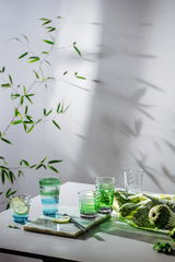 Ein Arrangement von Trinkgläsern in verschiedenen Grüntönen auf einem Tisch, umgeben von Pflanzen und einem Tuch, aus einer seitlichen Perspektive fotografiert.