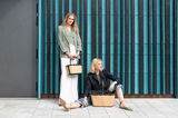 Zwei Frauen mit stilvollen, modernen Tragetaschen vor einer Wand mit vertikalen Streifen. Eine Frau steht und hält eine kleinere Tasche, während die andere sitzt und eine größere Tasche neben sich hat. Die Perspektive zeigt die Taschen von vorne.