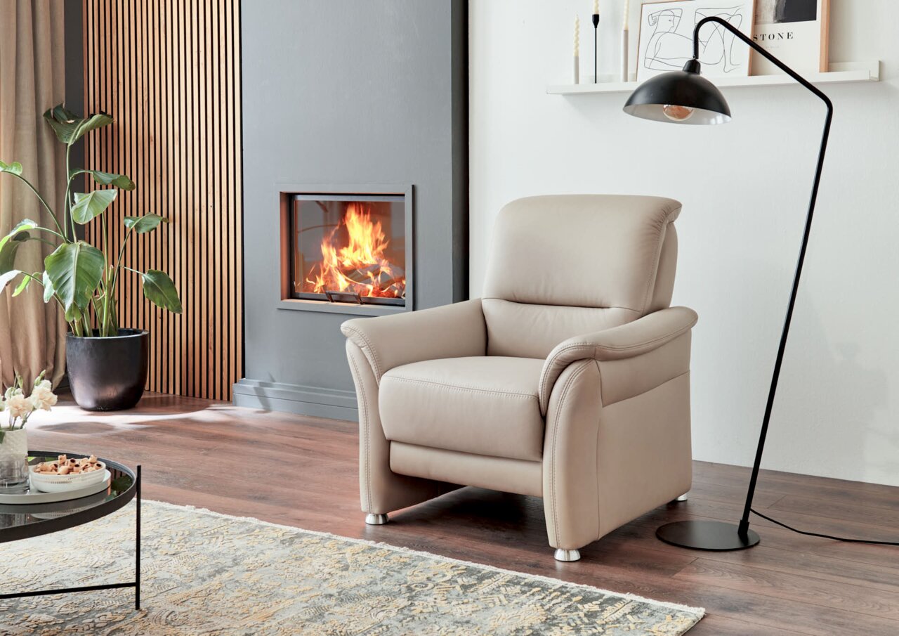Comfortabele beige fauteuil in een moderne woonkamer, gezien vanaf de zijkant, naast een open haard en een staande lamp.
