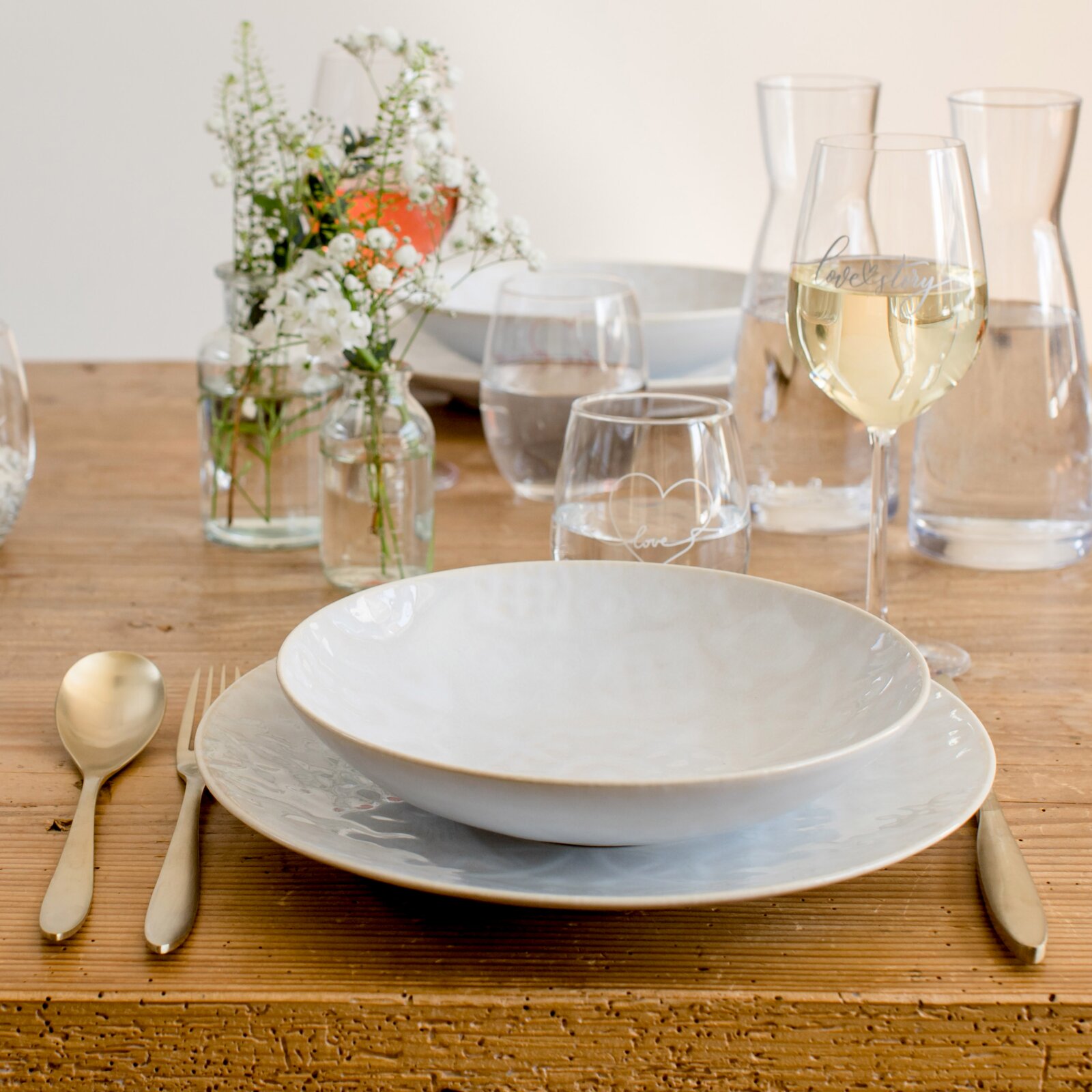 Gedeckter Holztisch mit weißem Geschirr, goldfarbenem Besteck, Wasserkaraffen und Gläsern sowie Blumen in Glasvasen, in leichter Schrägansicht von vorn