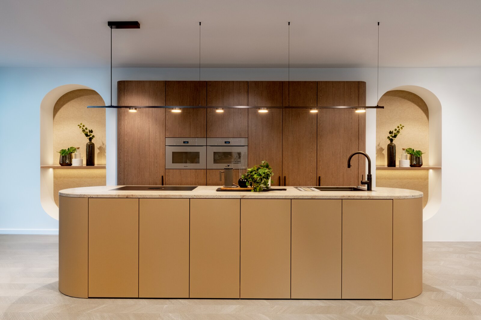 Lucide Aufhängeseil LINIAL Frontalansicht einer modernen Designküche mit großer Kücheninsel in Sandbeige und heller Steinplatte, dahinter Holzfronten mit Einbaugeräten, schwarzer Armatur und linearer Pendelleuchte sowie dekorativen Nischen mit Pflanzen.