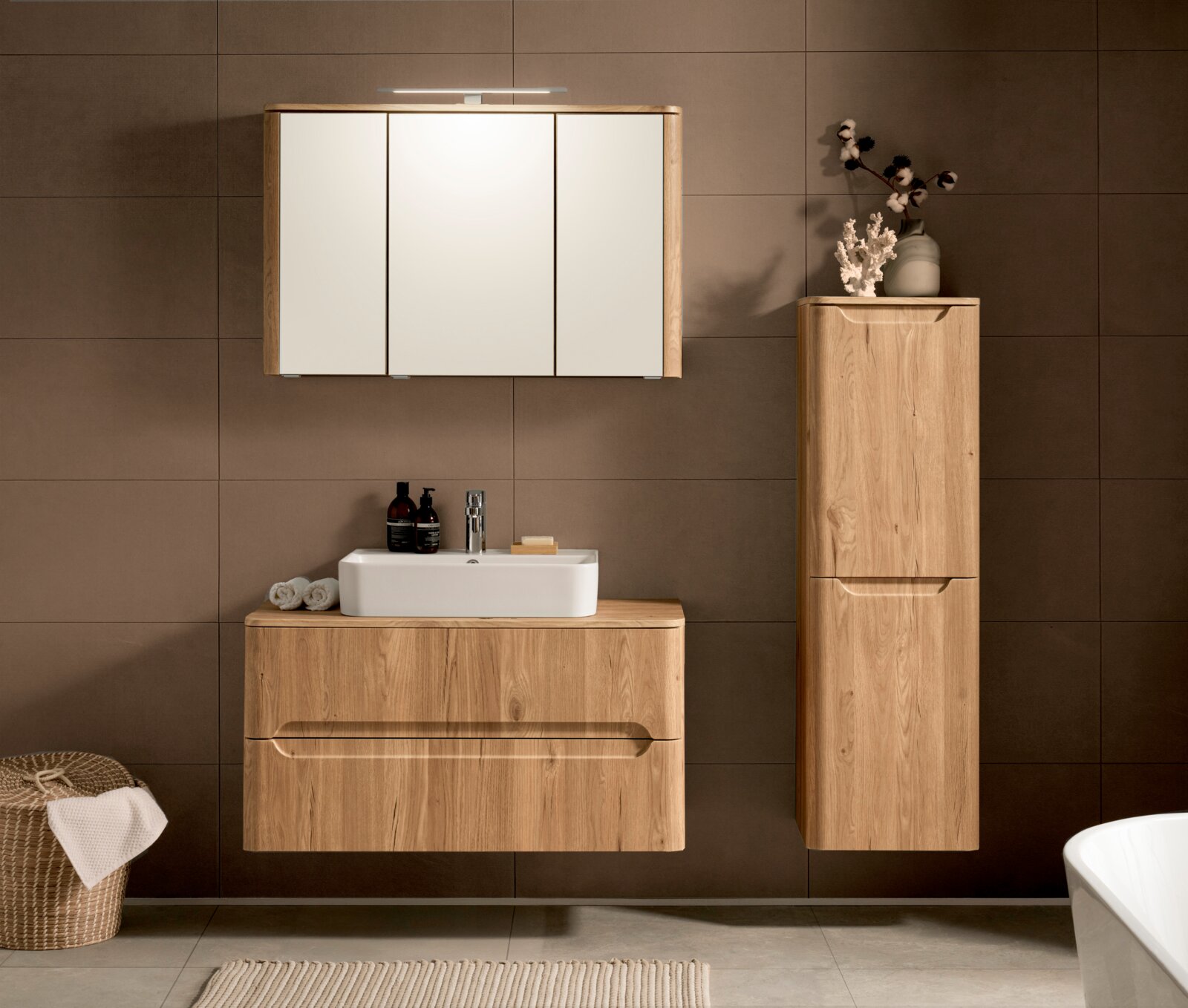 Modernes Badmöbel-Set in heller Holzoptik mit wandhängendem Waschtischunterschrank und weißem Aufsatzwaschbecken, Spiegelschrank mit drei Türen und LED-Leuchte sowie hohem Seitenschrank vor brauner Fliesenwand, Frontalansicht