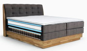 Boxspringbett mit grauem, gepolstertem Kopfteil und sichtbarem Schichtaufbau, seitliche Perspektive