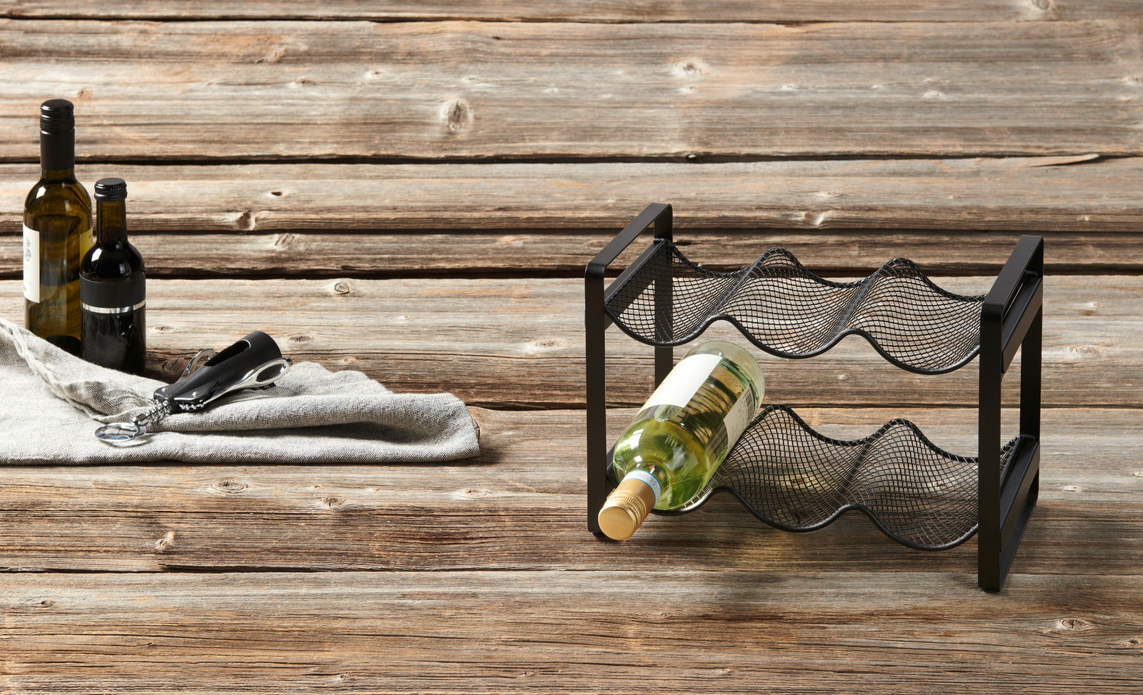 Ein schwarzes Flaschenregal aus Metall mit zwei Ebenen, auf einem Holztisch stehend. Eine Weinflasche liegt im Regal, daneben sind weitere Flaschen und ein Korkenzieher zu sehen. Perspektive von schräg oben.