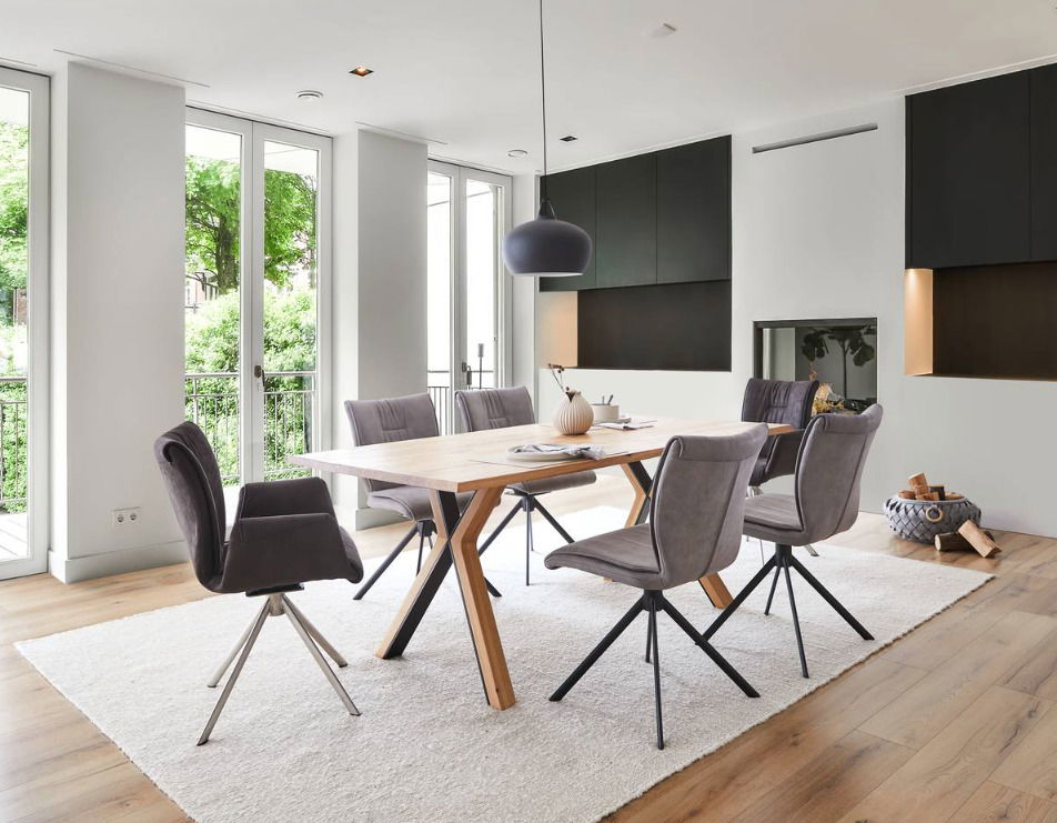 Moderner Esstisch mit Holztischplatte und schwarzen Metallbeinen, umgeben von grauen Stühlen in einem hellen Raum mit großen Fenstern, aus seitlicher Perspektive.