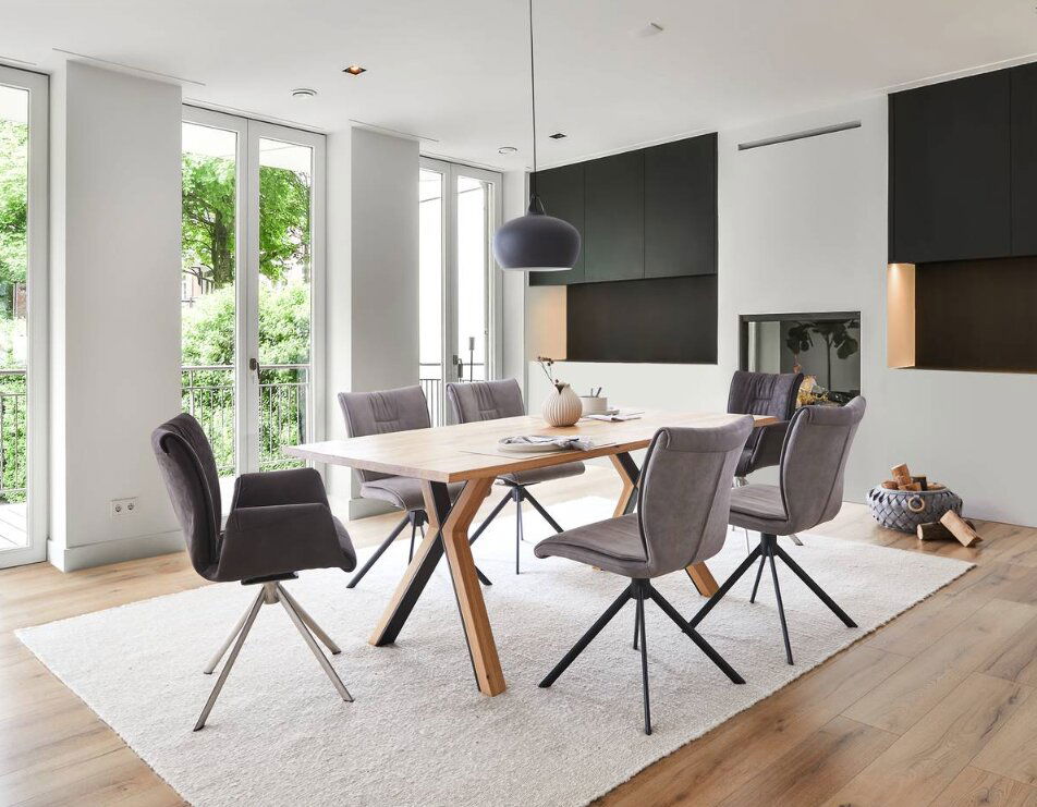 Moderner Esstisch mit Holztischplatte und schwarzen Metallbeinen, umgeben von grauen Stühlen in einem hellen Raum mit großen Fenstern, aus seitlicher Perspektive.