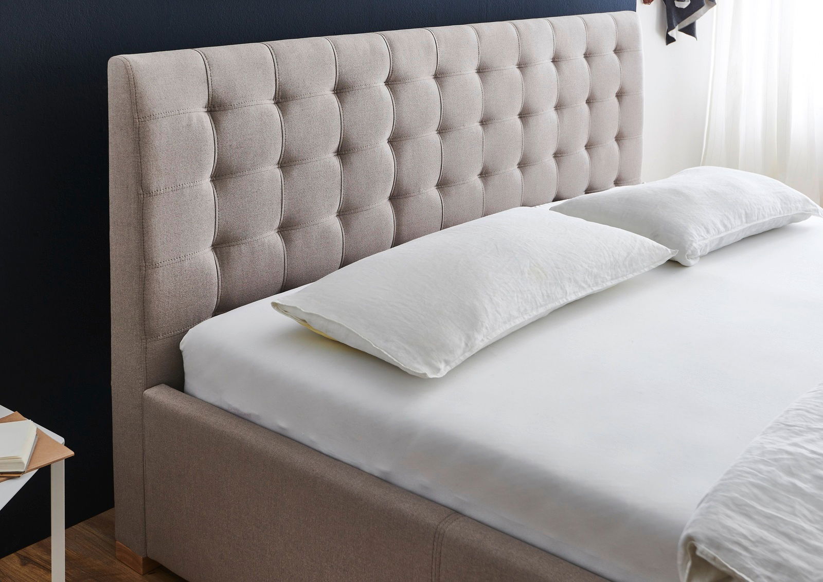 Polsterbett mit gestepptem Kopfteil in seitlicher Perspektive, bezogen mit beigem Stoff, aufgestellt in einem modernen Schlafzimmer.