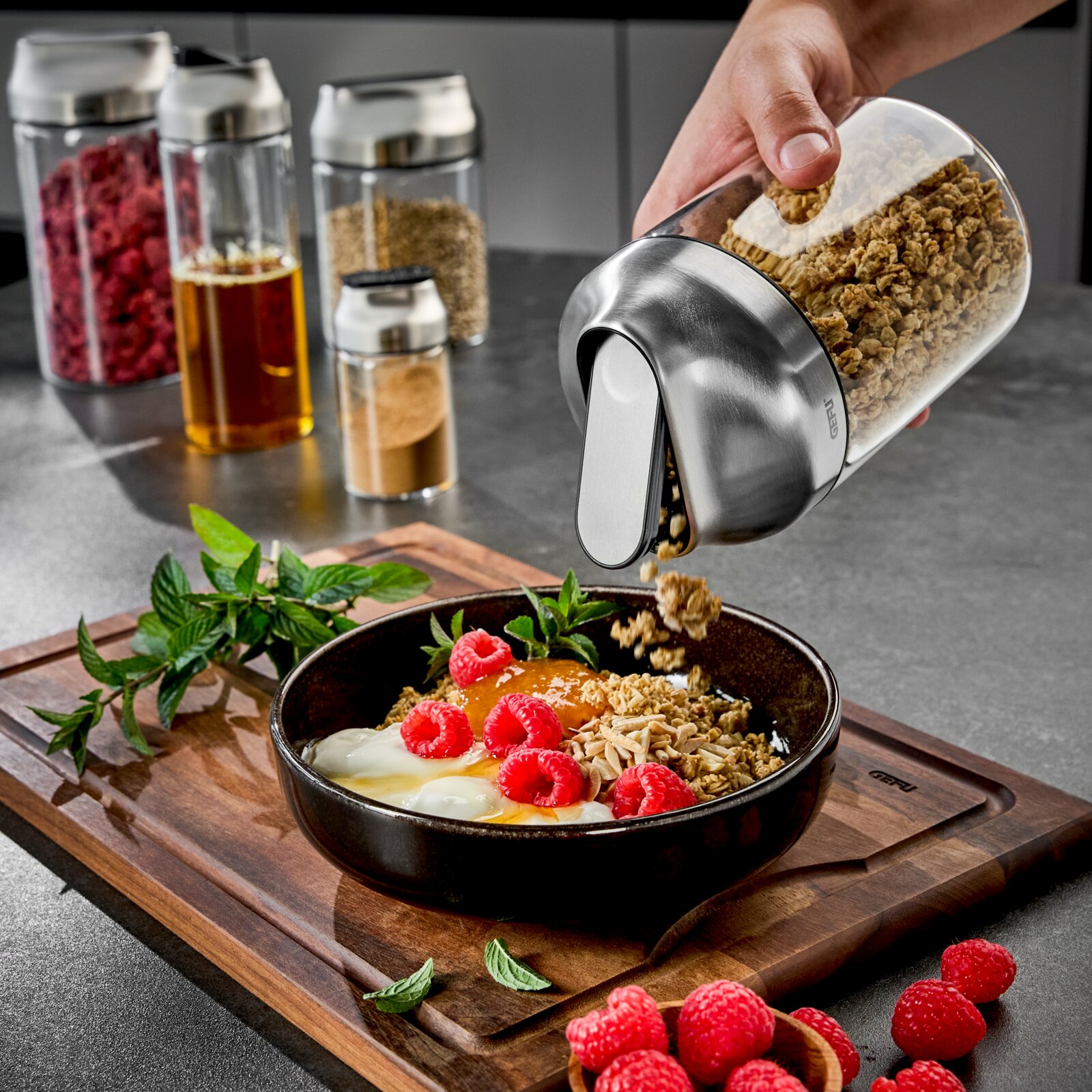 Hand hält eine Vorratsdose aus Glas mit Edelstahlverschluss, die Müsli in eine Schale mit Joghurt und Himbeeren auf einem Holzbrett schüttet. Im Hintergrund sind weitere Vorratsdosen zu sehen. Perspektive: seitlich.