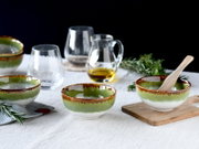Grün-weiß glasierte Keramikschalen mit braunem Rand auf hellem Leinentuch, daneben Glas-Karaffe mit Öl, Trinkgläser, Holzbrett und Kräuterzweige; fotografiert in leicht schräger Frontansicht auf Tischhöhe.