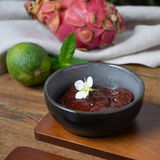 Anthrazitfarbene Dip-Schale mit roter Sauce und weißer Blüte, umgeben von einer Limette und einer Drachenfrucht, aus der Vogelperspektive.