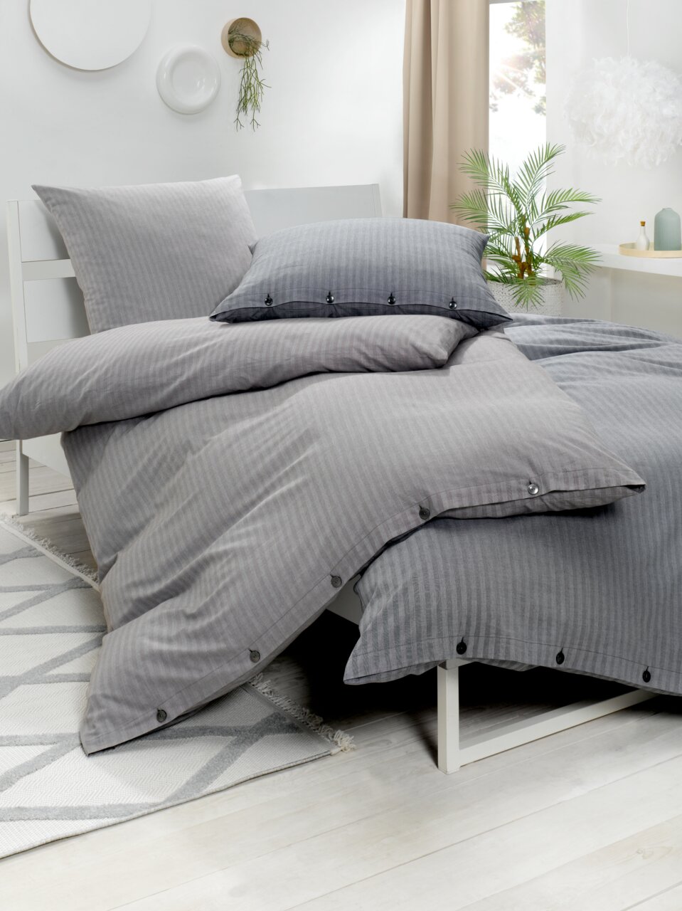 Zurbrüggen Bettwäsche 135x200cm + 80x80cm HERINGBONE Graue Bettwäsche im Heringbone-Muster auf einem weißen Bett, seitliche Perspektive mit dekorativen Kissen und Pflanzen im Hintergrund.