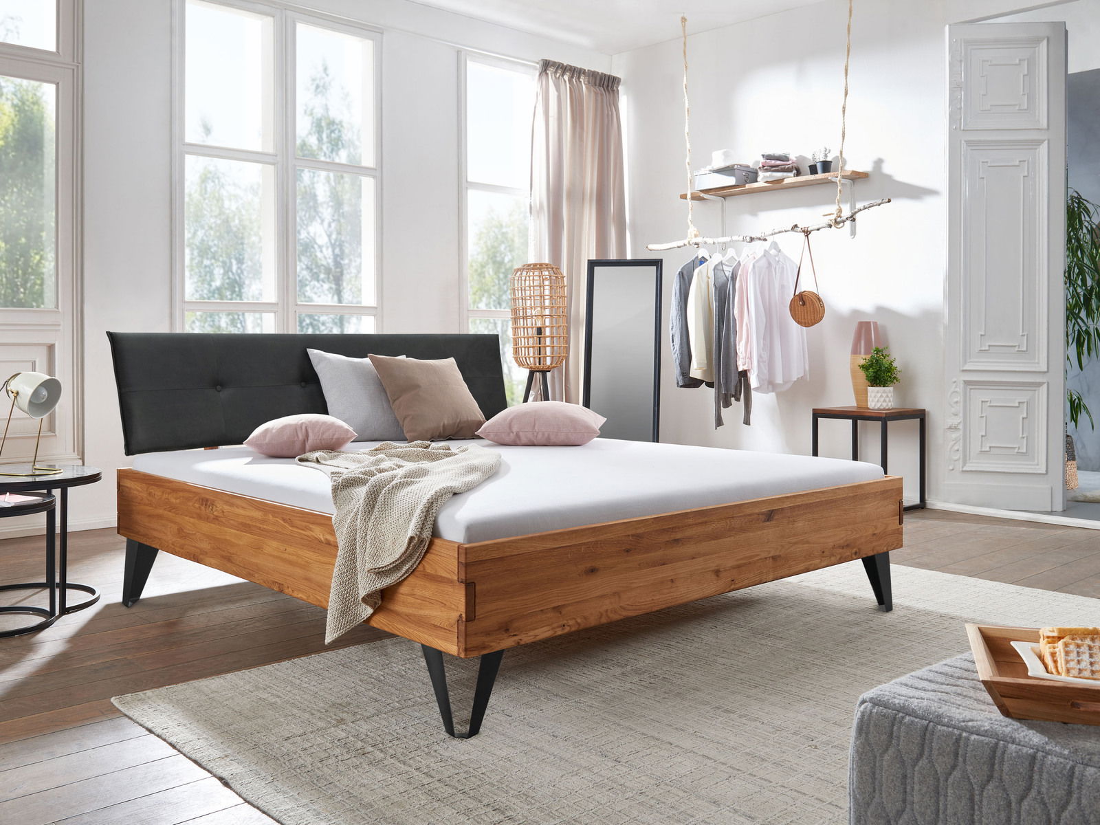 Modernes Bett aus Holz mit schwarzem Kopfteil, seitliche Perspektive, in einem hellen Schlafzimmer mit großen Fenstern und stilvoller Dekoration.