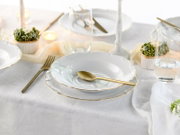 Elegantes weißes Porzellan-Tafelservice mit goldfarbenem Rand (Teller und Suppenteller) mit goldenem Besteck und Wassergläsern auf weißer Tischdecke, perspektivisch in leicht schräger Seitenansicht von vorne fotografiert.