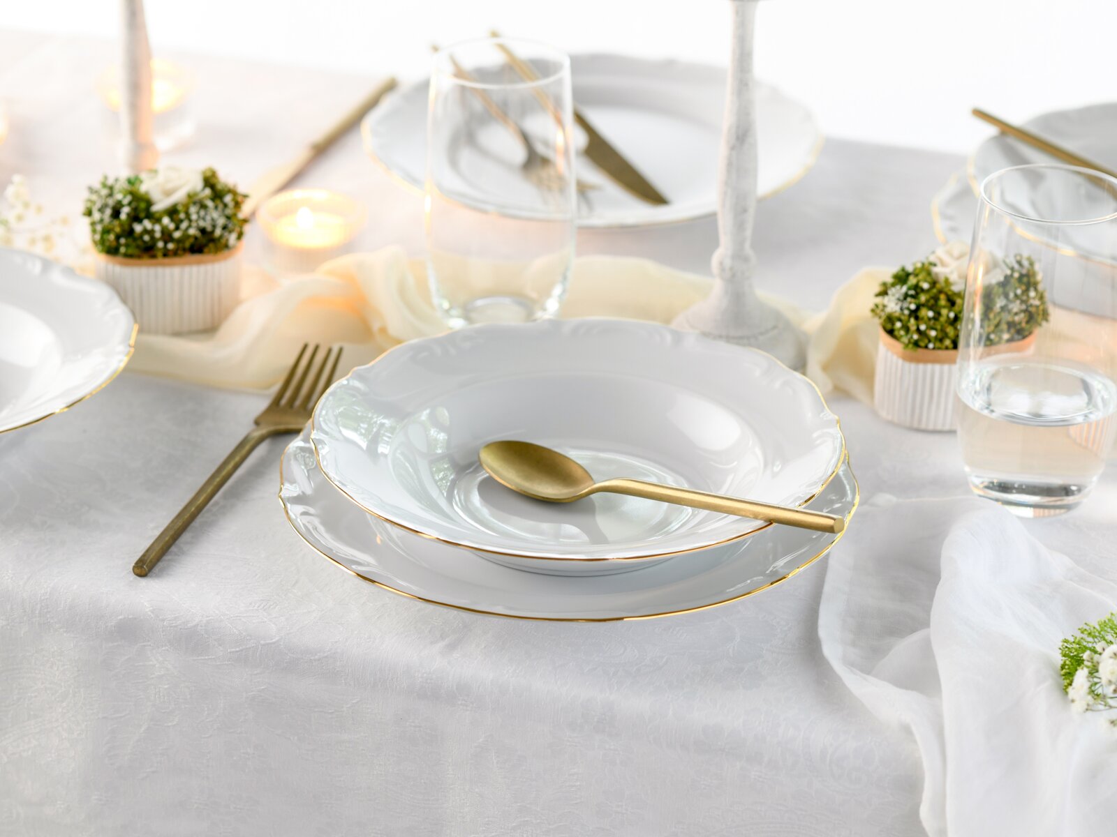 Elegantes weißes Porzellan-Tafelservice mit goldfarbenem Rand (Teller und Suppenteller) mit goldenem Besteck und Wassergläsern auf weißer Tischdecke, perspektivisch in leicht schräger Seitenansicht von vorne fotografiert.