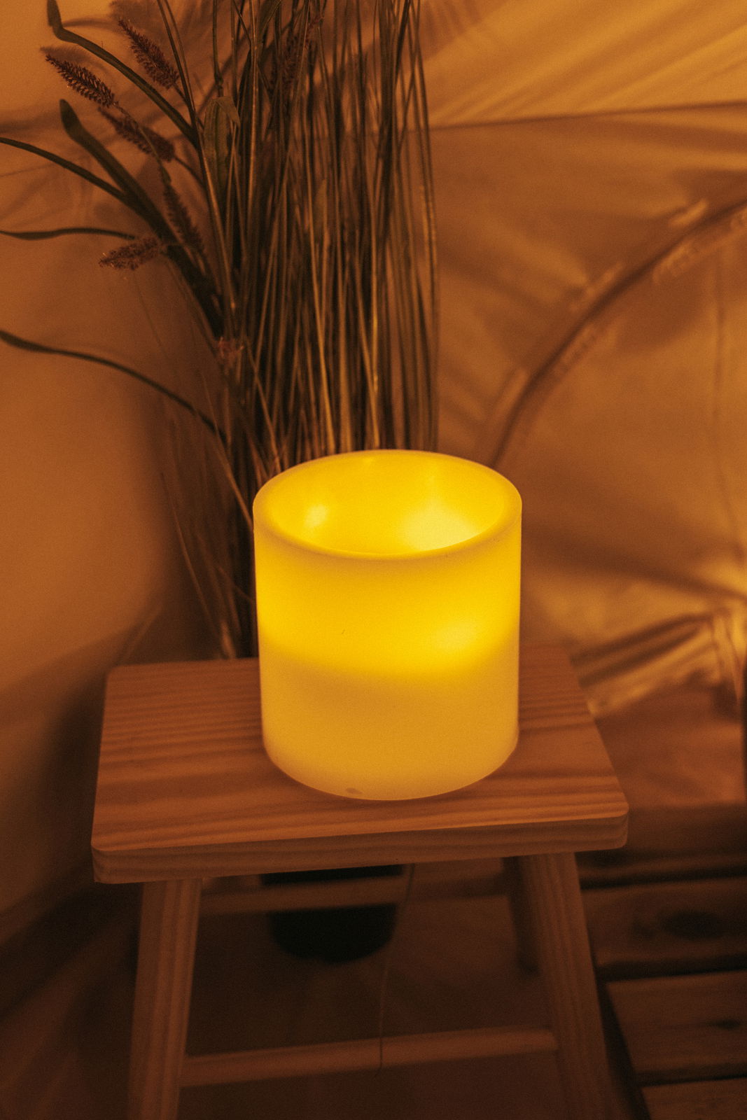 Leuchtende Outdoor-Kerze auf einem Holzhocker, umgeben von Pflanzen, aus seitlicher Perspektive fotografiert.
