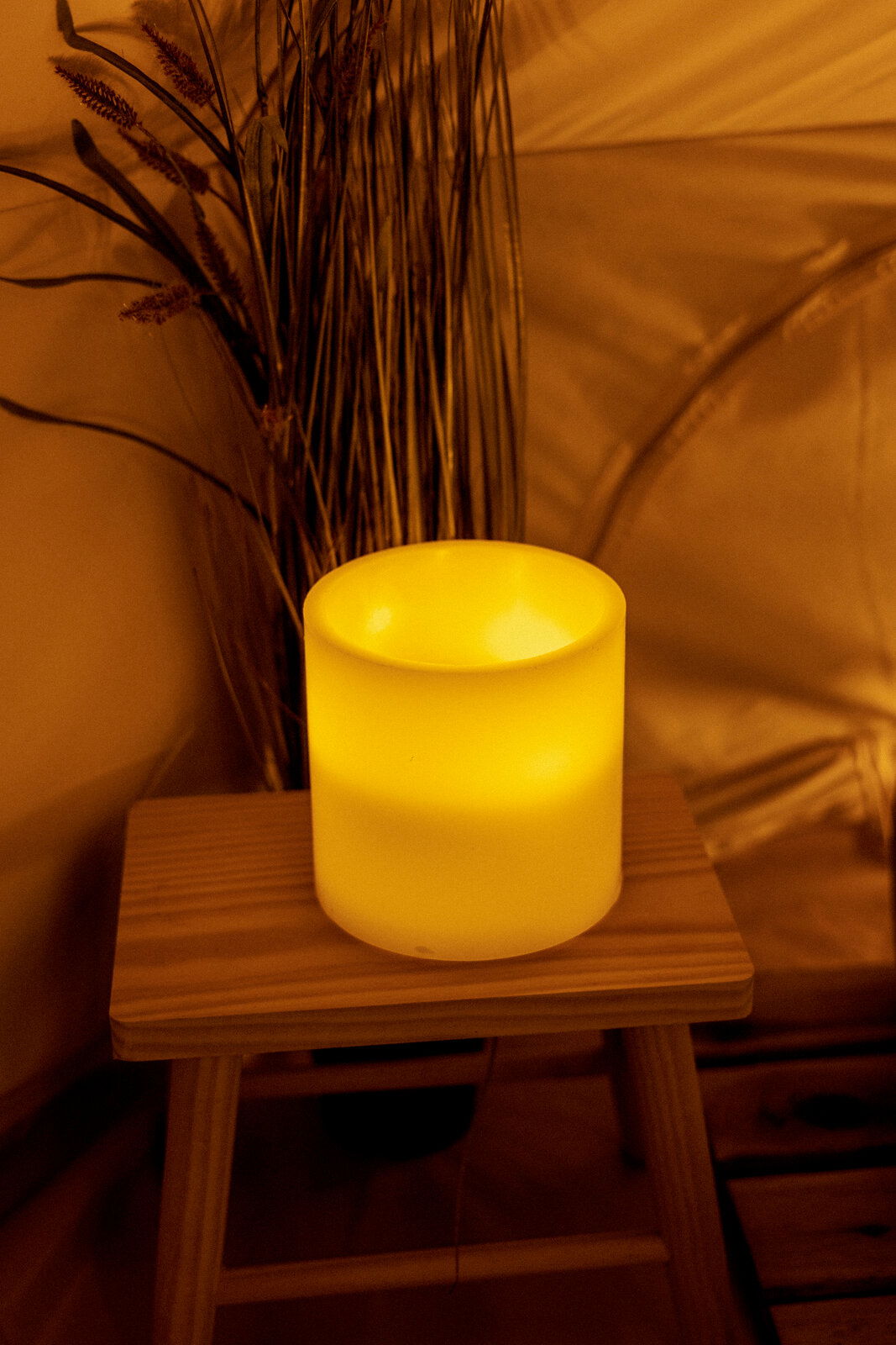 Leuchtende Outdoor-Kerze auf einem Holzhocker, umgeben von Pflanzen, aus seitlicher Perspektive fotografiert.