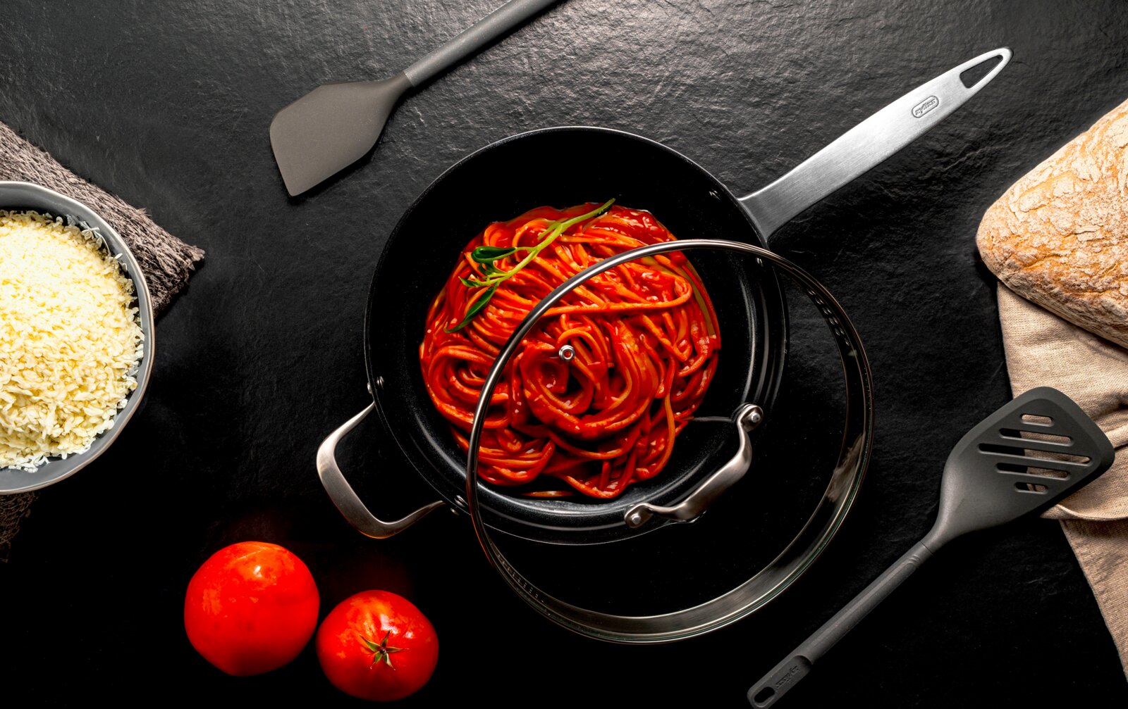Draufsicht auf einen schwarzen Kochtopf mit Glasdeckel und Spaghetti in Tomatensauce, umgeben von Küchenutensilien, Tomaten, Reis und Brot auf dunklem Hintergrund.