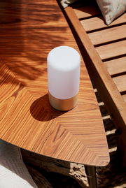 Kompakte, zylindrische Tischleuchte mit weißem Diffusor und goldfarbenem Sockel auf einem Holz-Beistelltisch im Sonnenlicht, schräg von oben fotografiert.