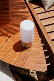 Kompakte, zylindrische Tischleuchte mit weißem Diffusor und goldfarbenem Sockel auf einem Holz-Beistelltisch im Sonnenlicht, schräg von oben fotografiert.