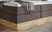 Nahaufnahme eines Boxspringbetts mit braunem Stoffbezug und hellem Holzrahmen, von der Seite betrachtet.
