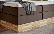 Nahaufnahme eines Boxspringbetts mit braunem Stoffbezug und hellem Holzrahmen, von der Seite betrachtet.