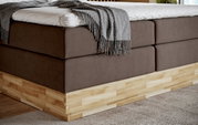 Close-up van een boxspringbed met een bruine stoffen bekleding en een licht houten frame, gezien vanaf de zijkant.