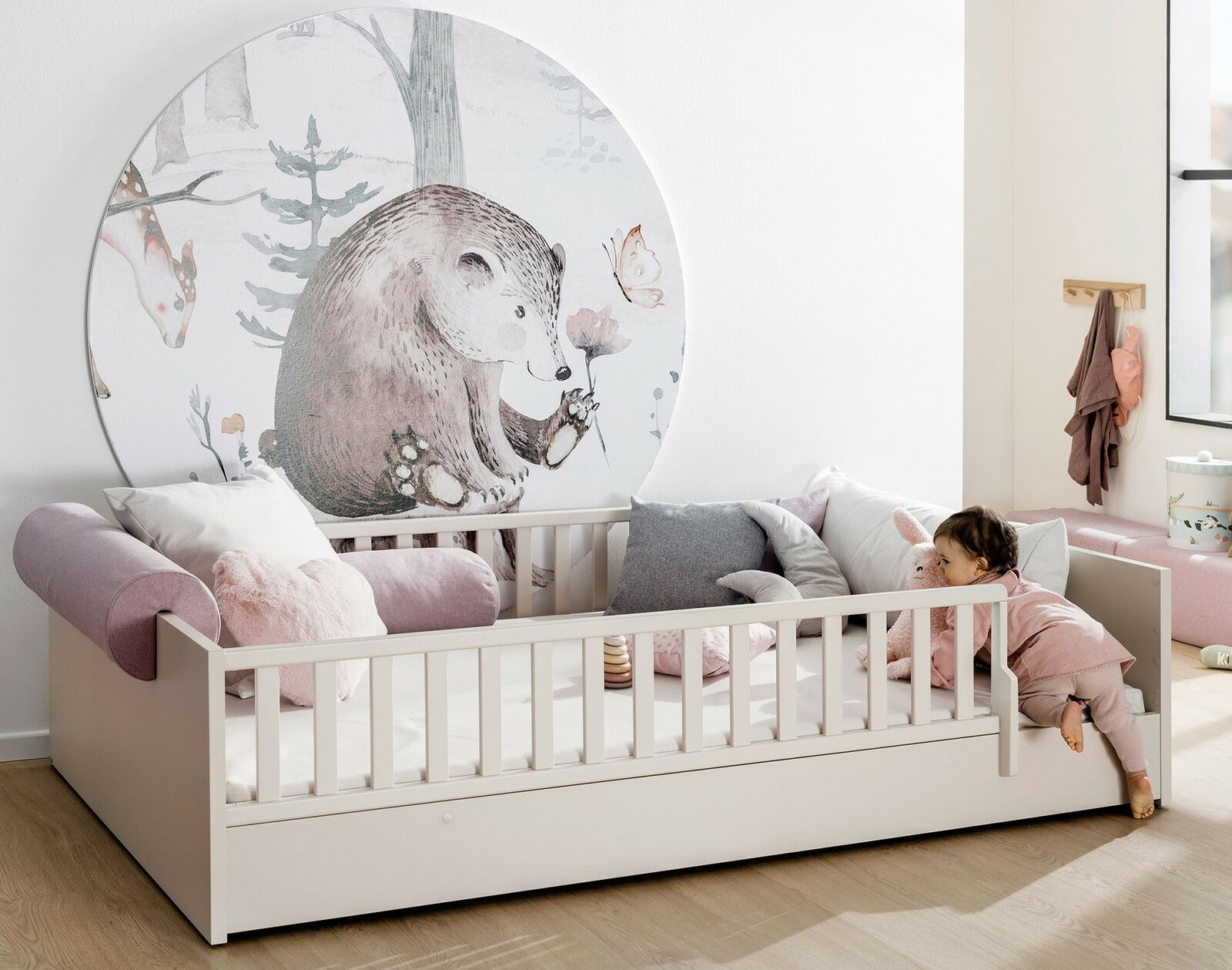 Kinderbett mit Rausfallschutz und Bettkasten, dekoriert mit Kissen und einem Wandbild eines Bären, seitliche Perspektive