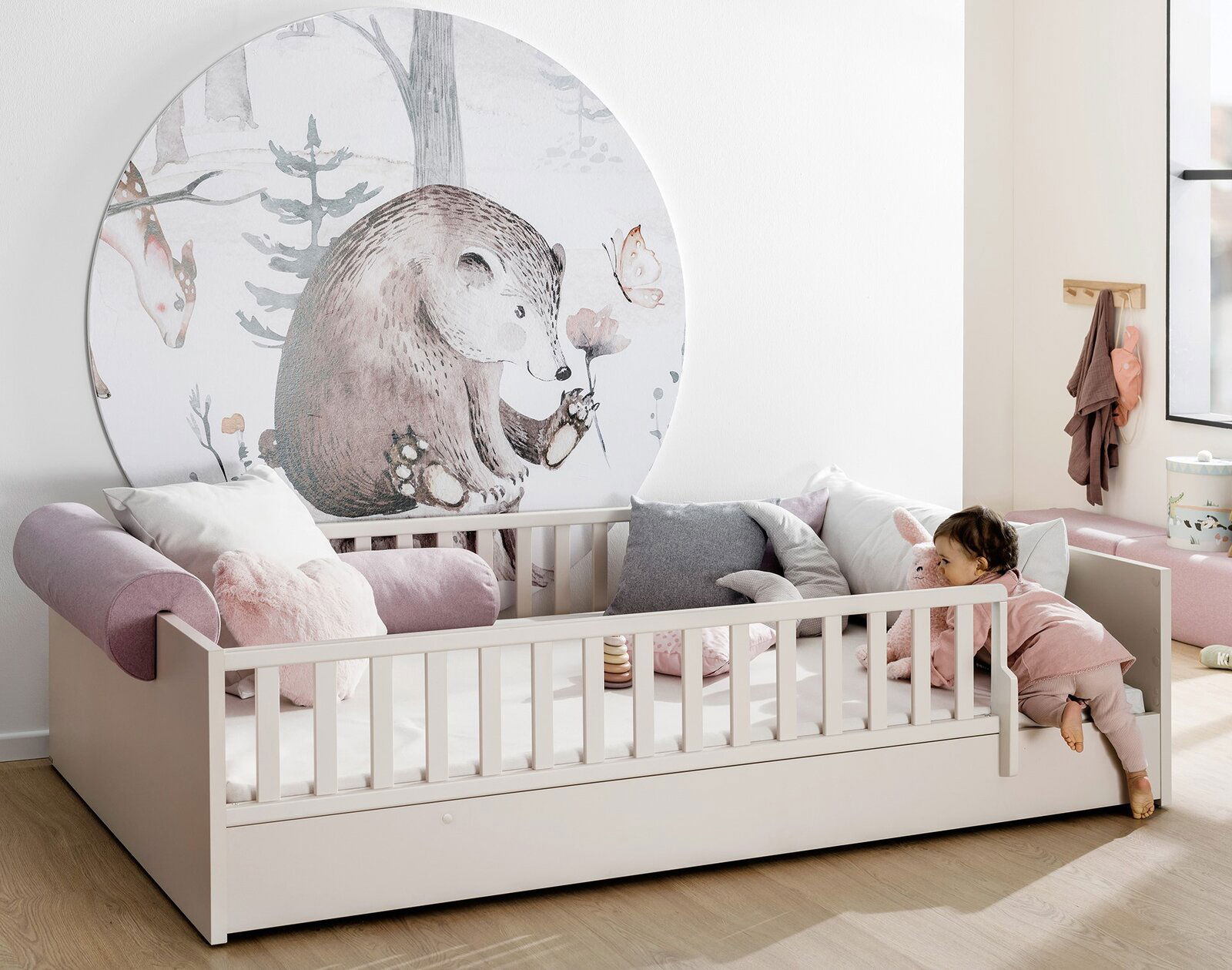 Kinderbett mit Rausfallschutz und Bettkasten, dekoriert mit Kissen und einem Wandbild eines Bären, seitliche Perspektive