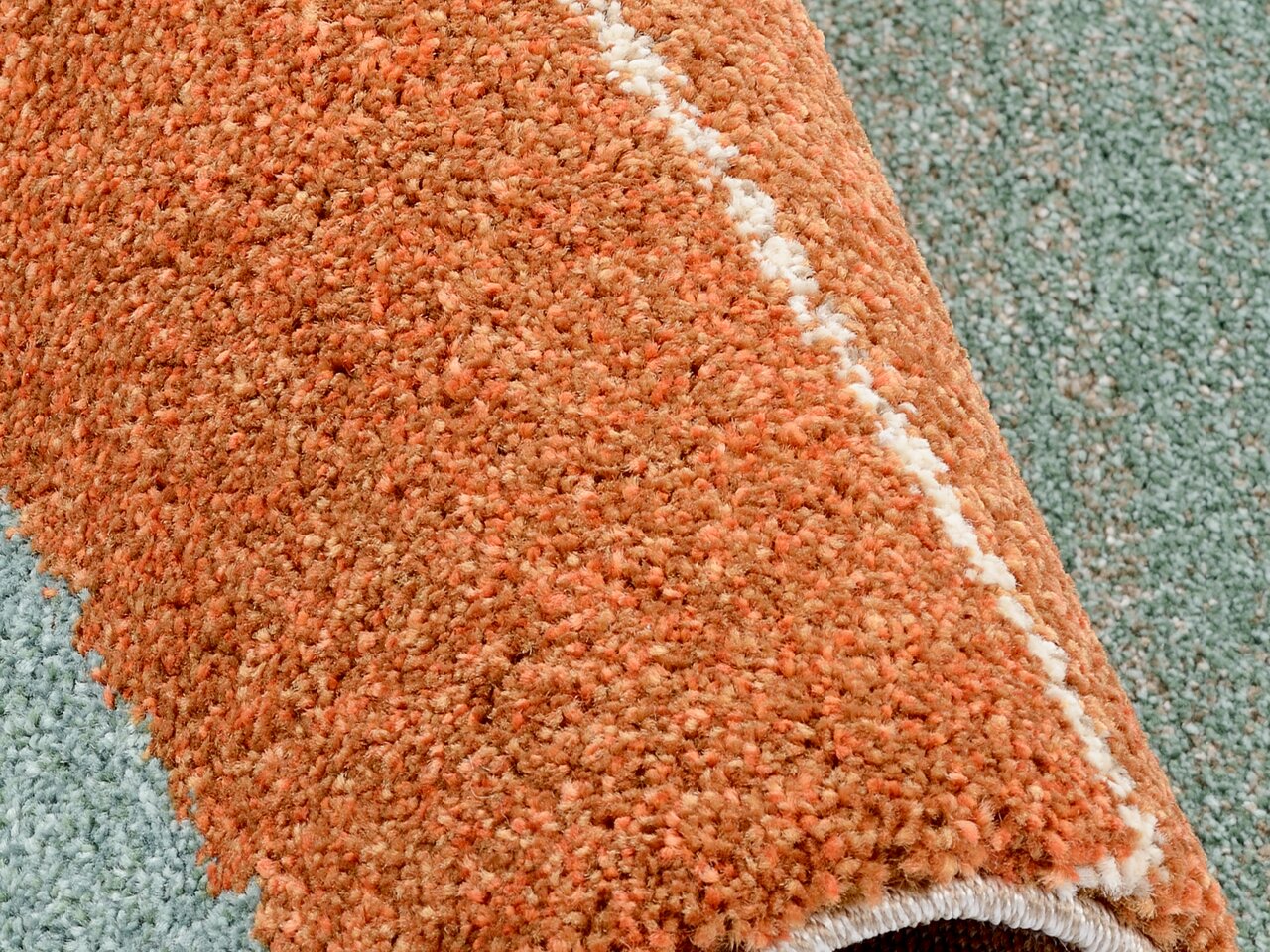 Nahaufnahme eines orangefarbenen Frisé-Teppichs mit einer weißen Linie, teilweise aufgerollt auf einem grünlichen Hintergrund.