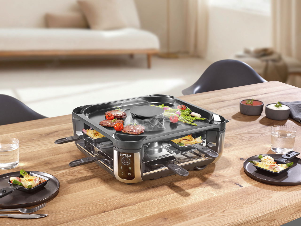 Smokeless Raclette-Grill auf einem Tisch, von schräg oben fotografiert, mit Grillgut und Pfännchen.