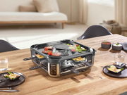 Smokeless Raclette-Grill auf einem Tisch, von schräg oben fotografiert, mit Grillgut und Pfännchen.