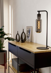 Moderne Tischleuchte MAIONE mit schwarzem Gestell und dekorativer Glühbirne, seitliche Perspektive auf einem Holztisch.