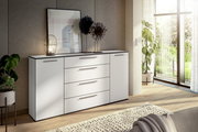 Modernes weißes Sideboard mit Schubladen und Türen, dekoriert mit einer Lampe und Kunstobjekten, in einem hellen Raum mit Blick auf den Garten. Perspektive von schräg vorne.