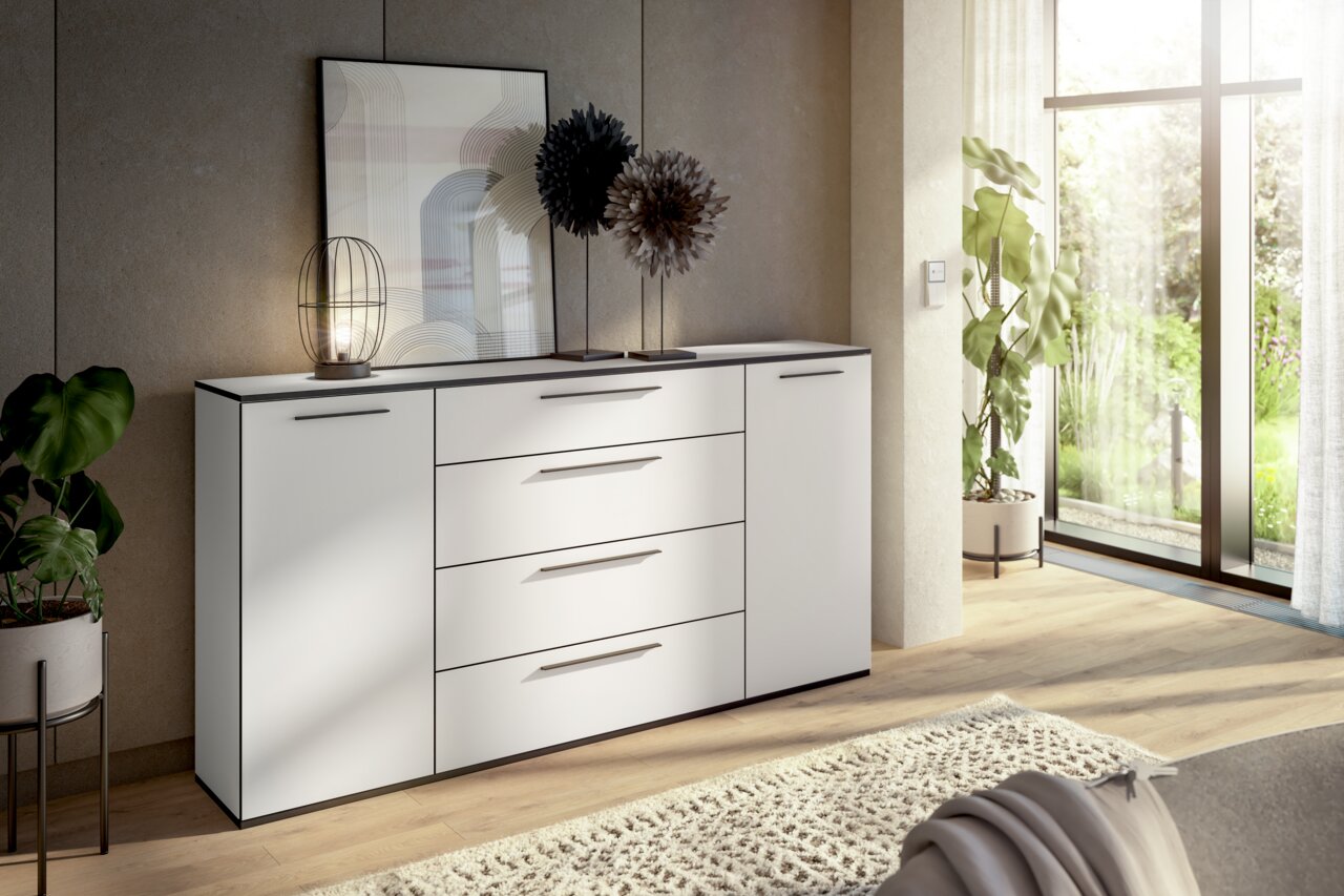 Modernes weißes Sideboard mit Schubladen und Türen, dekoriert mit einer Lampe und Kunstobjekten, in einem hellen Raum mit Blick auf den Garten. Perspektive von schräg vorne.