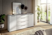 Modernes weißes Sideboard mit Schubladen und Türen, dekoriert mit einer Lampe und Kunstobjekten, in einem hellen Raum mit Blick auf den Garten. Perspektive von schräg vorne.