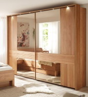Großer Kleiderschrank in Holzoptik mit drei Schiebetüren, zwei davon verspiegelt, im hellen Schlafzimmer; Frontansicht leicht schräg von links