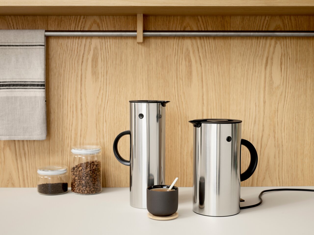 Zwei Edelstahl-Wasserkocher EM77 mit schwarzem Griff und Deckel, auf einer Küchenarbeitsplatte vor einer Holzwand, aus seitlicher Perspektive.