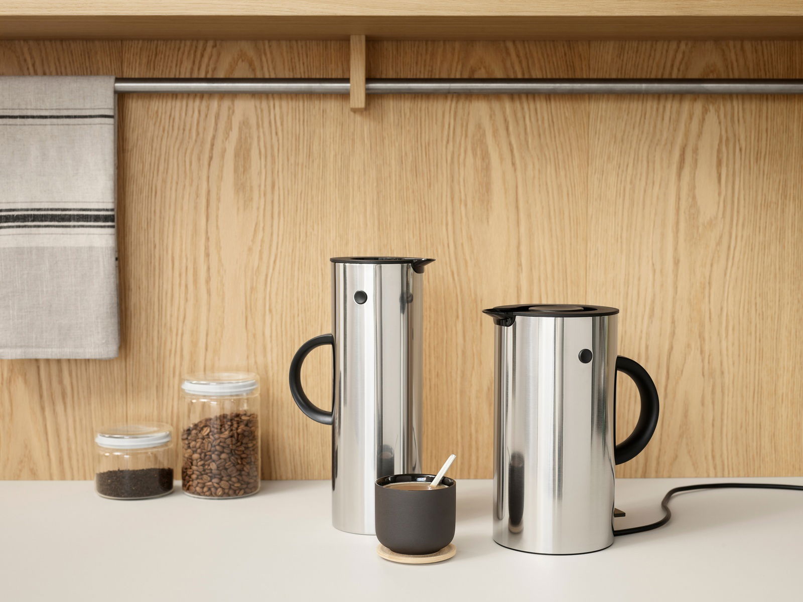 Zwei Edelstahl-Wasserkocher EM77 mit schwarzem Griff und Deckel, auf einer Küchenarbeitsplatte vor einer Holzwand, aus seitlicher Perspektive.