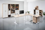 Moderne Büromöbel in einem hellen Raum, bestehend aus einem L-förmigen Schreibtisch mit Holzoberfläche und weißen Beinen, einem Rollcontainer und mehreren Regalen mit Schubladen und Türen. Perspektive von vorne.