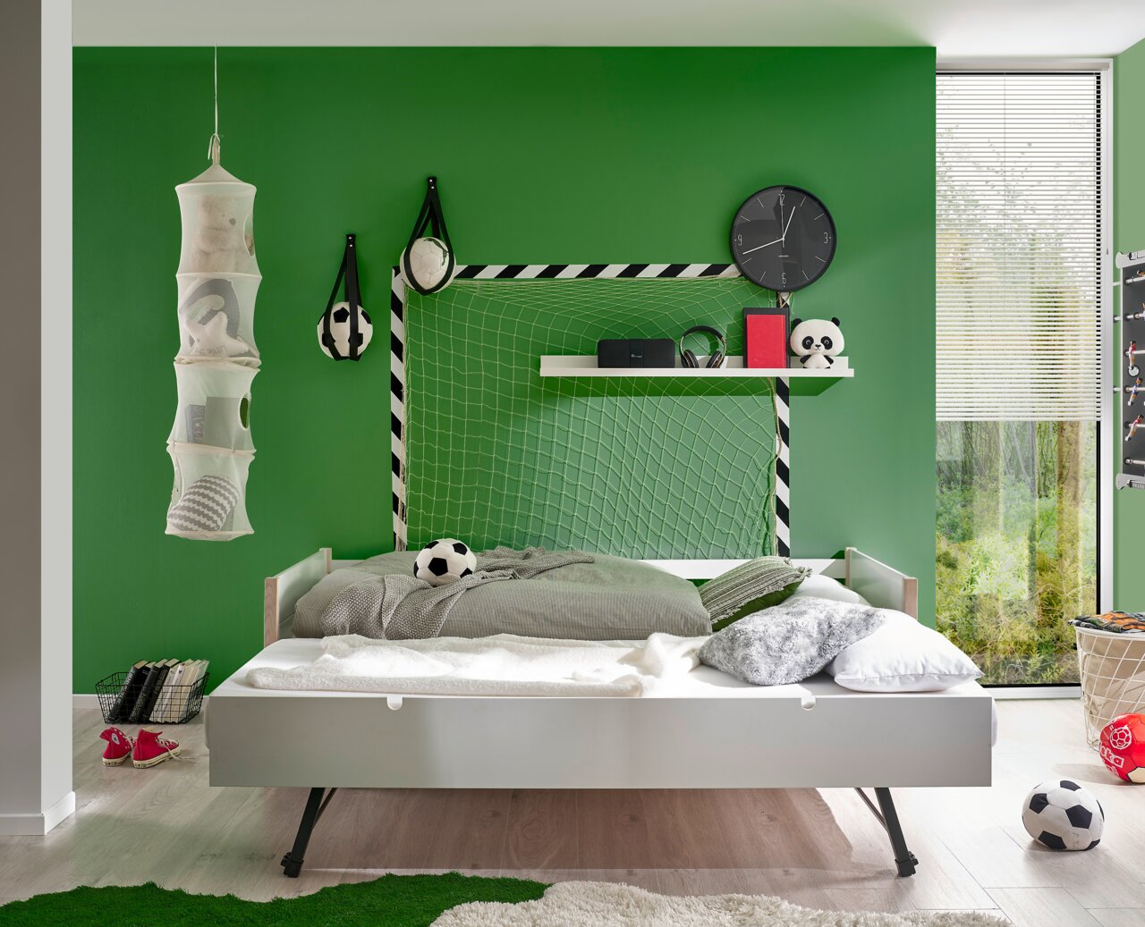 Jugendbett in einem Kinderzimmer mit grün gestrichener Wand, Fußball-Dekorationen und einem Regal. Perspektive von vorne.