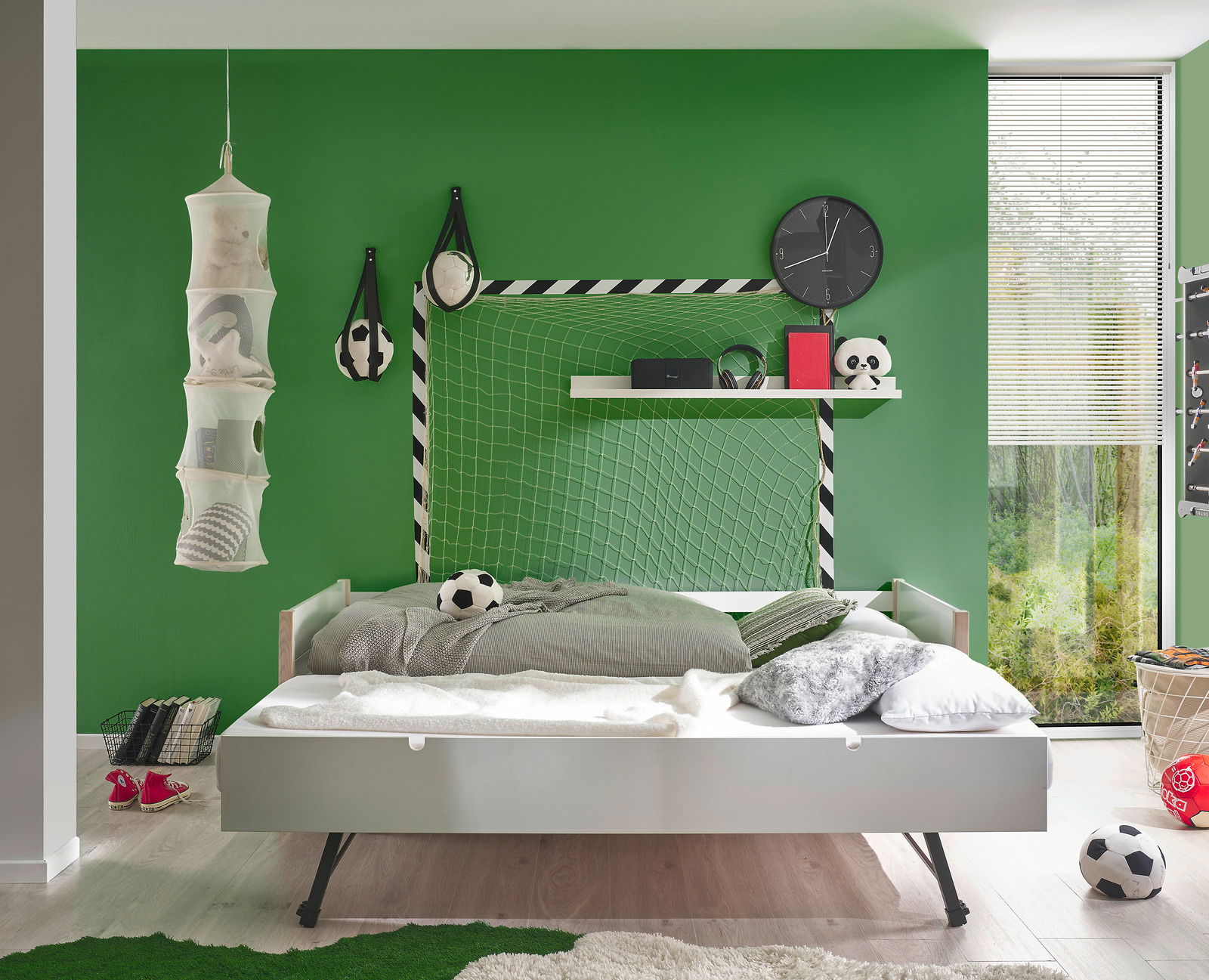 Jugendbett in einem Kinderzimmer mit grün gestrichener Wand, Fußball-Dekorationen und einem Regal. Perspektive von vorne.