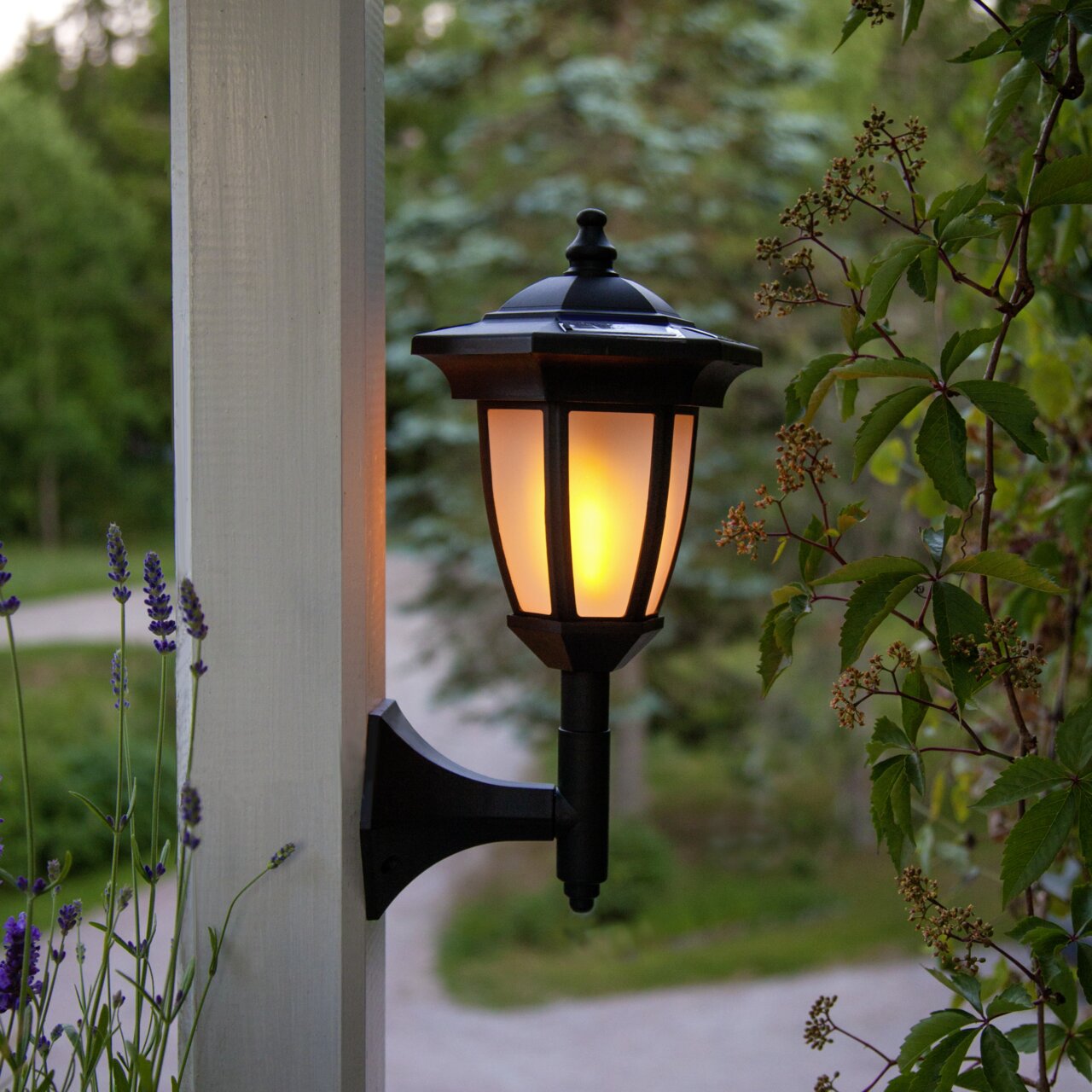 STAR TRADING LED-Solar-Laterne 4 in 1 FLAME 4 IN 1 Schwarze Außen-Wandleuchte im Laternenstil mit warmem Licht, seitliche Ansicht an einem weißen Pfosten im Garten