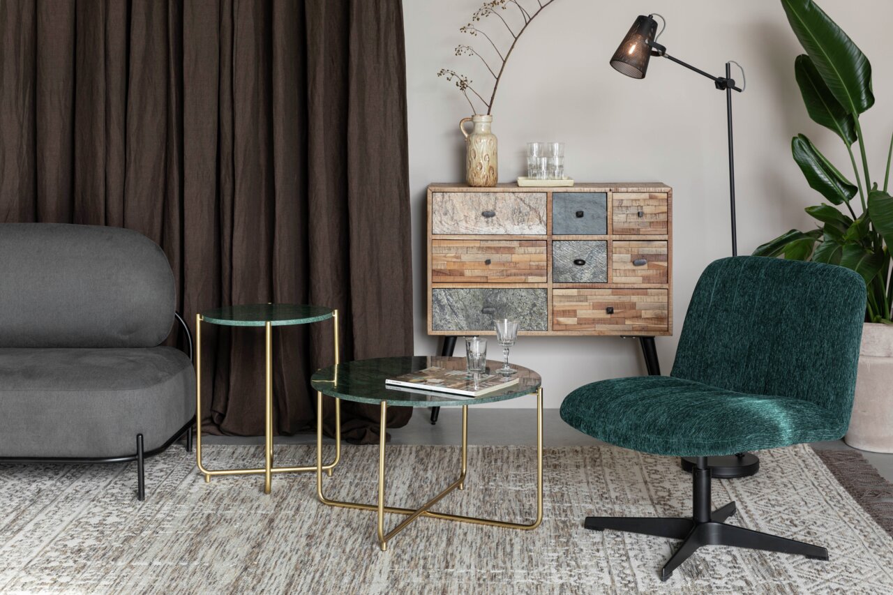 Een moderne woonkamer met een groene BELMONDO-fauteuil die vanaf de voorkant zichtbaar is, naast een ronde tafel met gouden poten en een dressoir op de achtergrond.