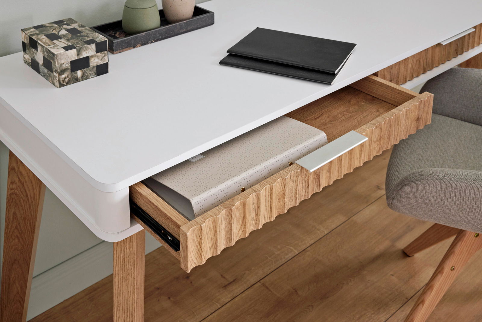 Moderner Schreibtisch mit weißer Oberfläche und Holzbeinen, aus der Perspektive von oben fotografiert. Eine Schublade ist teilweise geöffnet und zeigt darin liegende Ordner.