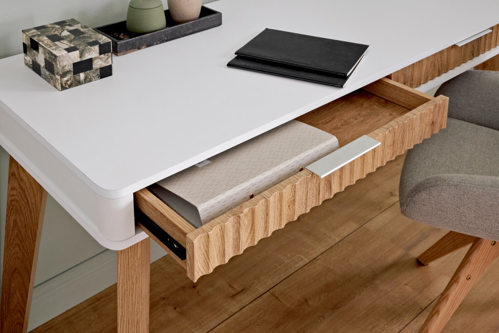 Moderner Schreibtisch mit weißer Oberfläche und Holzbeinen, aus der Perspektive von oben fotografiert. Eine Schublade ist teilweise geöffnet und zeigt darin liegende Ordner.