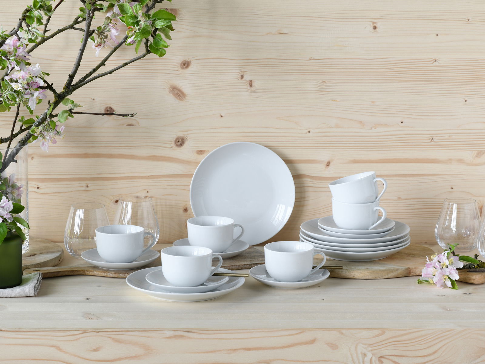 Kaffeeservice 18-teilig in eleganter Anordnung auf einem Holztisch, mit weißen Tassen und Tellern, umgeben von blühenden Zweigen, aus frontaler Perspektive.