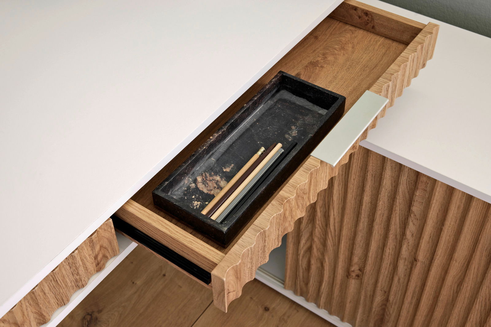 Nahaufnahme eines Eck-Schreibtisches mit Anbau, der eine geöffnete Schublade mit einem schwarzen Tablett und Stiften zeigt. Die Perspektive ist von oben.