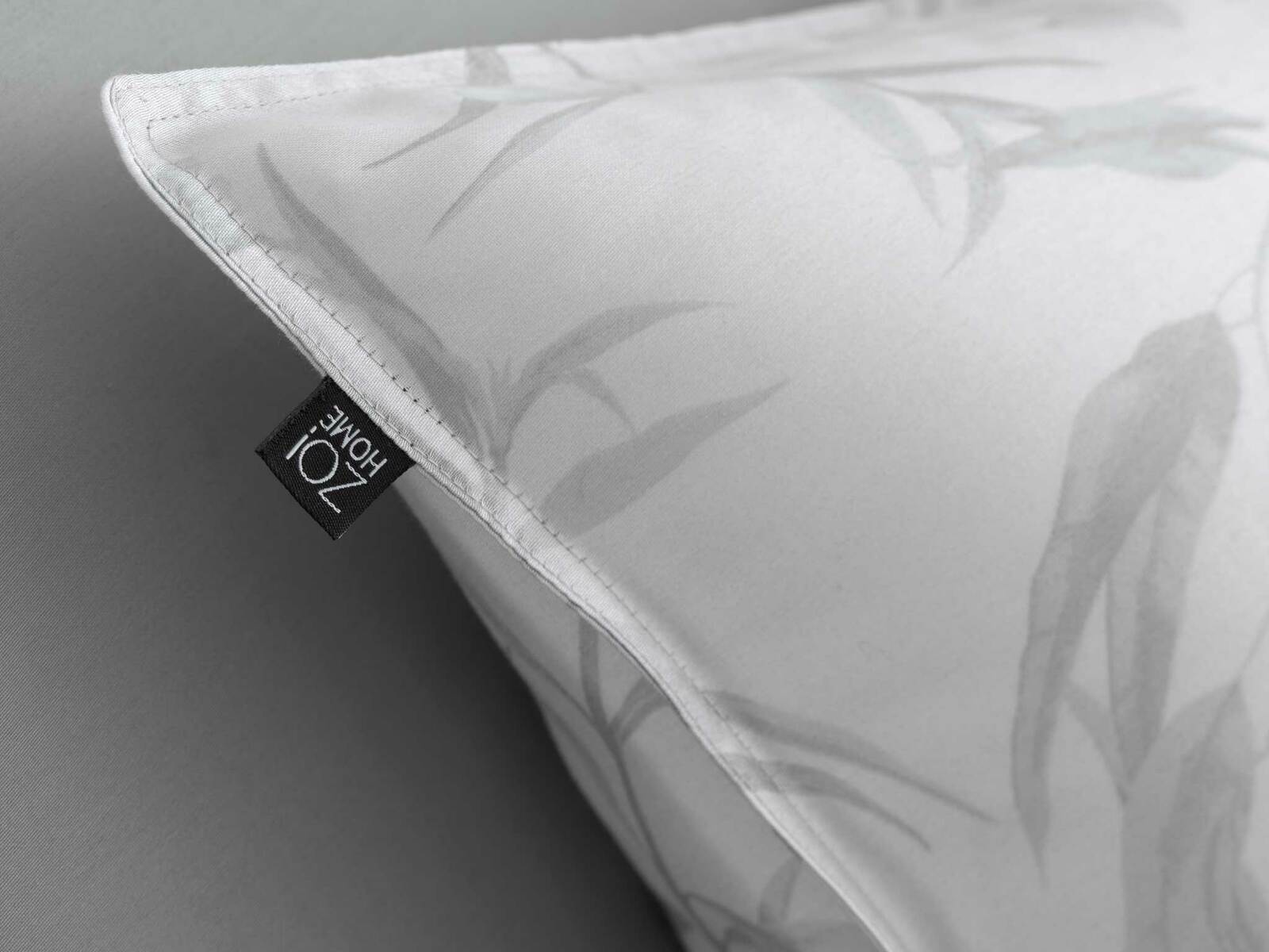 Nahaufnahme eines weißen Kissenbezugs mit dezentem Blattmuster und einem Zoi Home Etikett an der Ecke.