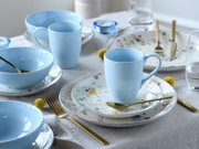 Gedeckter Esstisch mit hellblauen Keramikbechern und -schalen sowie weißem, bunt gesprenkeltem Geschirr und goldfarbenem Besteck, fotografiert in schräger Draufsicht (3/4-Perspektive)